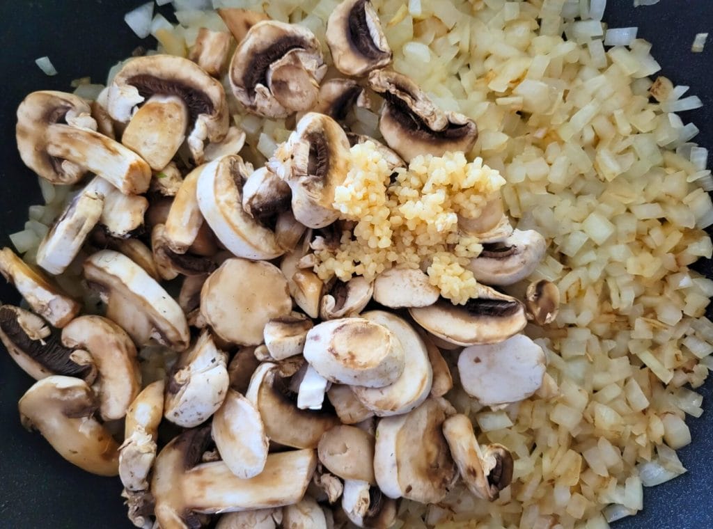 sauteed, diced onions in a black skillet with sliced mushrooms and minced garlic on top