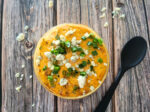 jackfruit buffalo dip in a clear bowl, sprinkled with sliced green onions and blue cheese, on a wooden surface, next to a black mixing spoon