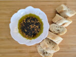 olive oil bread dip in a white bowl stylized like a flower on a light wooden surface, next to a sliced baguette.
