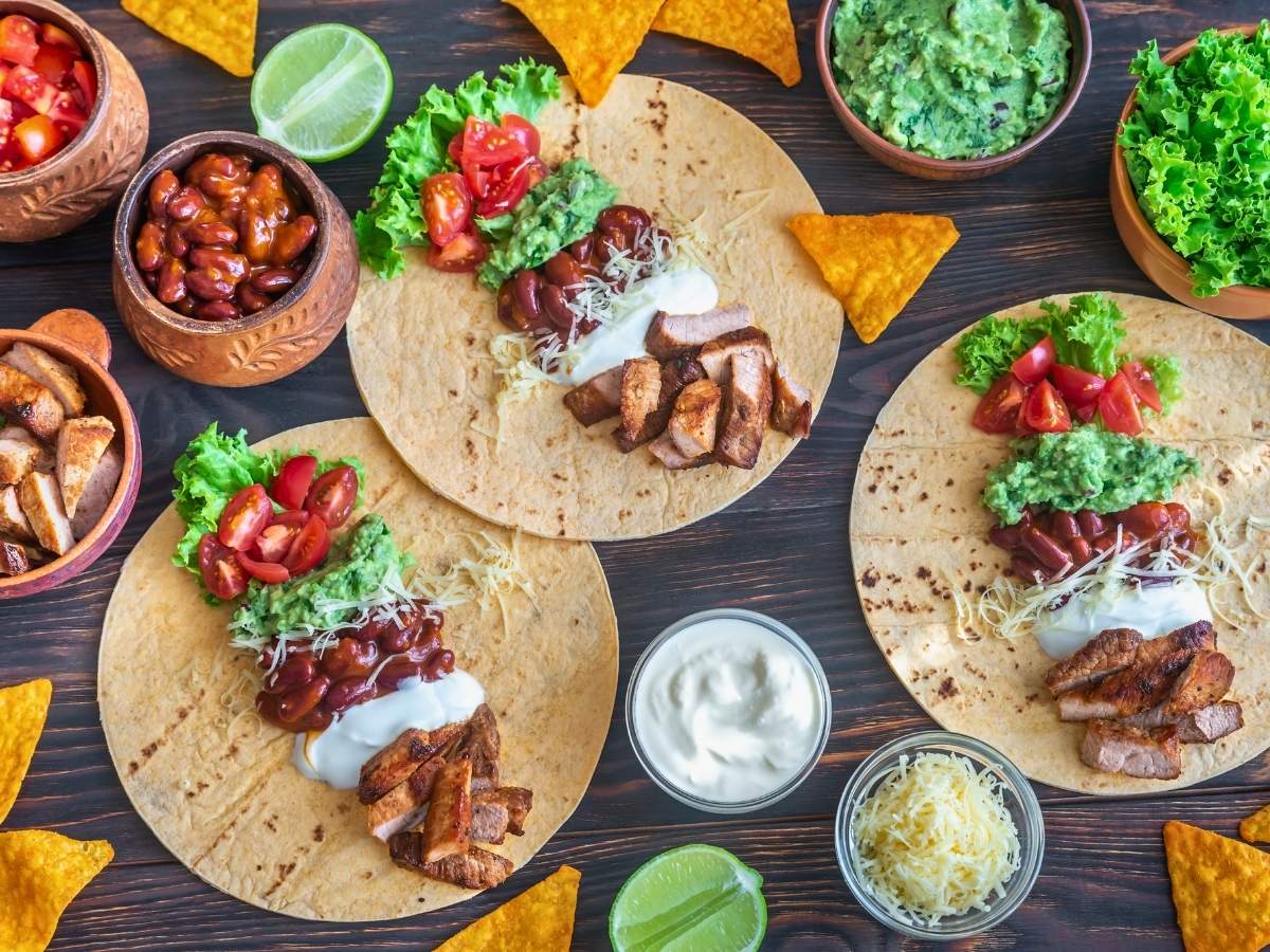 3 flour tortillas on a grey wooden surface. the have layers of taco fillings on them, including chicken, beans, sour cream, tomatoes, and mashed avocado on them. They are surrounded by various taco fillings in individual bowls.
