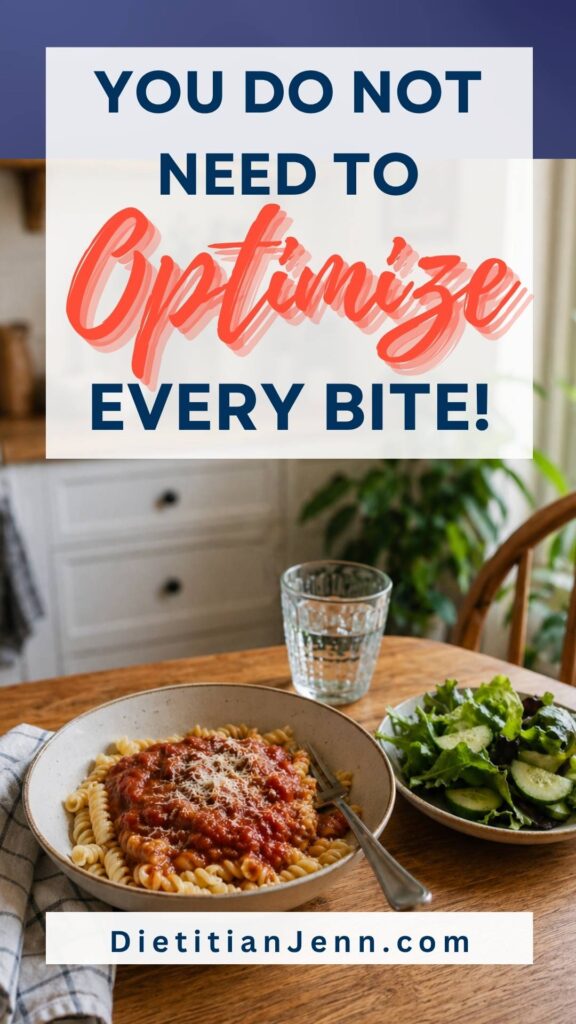 A bowl of rotini pasta with marinara sauce and grated cheese, next to a small salad and a glass of water on a wooden table. text above reads, "You Do Not Need to Optimize Every Bite!"